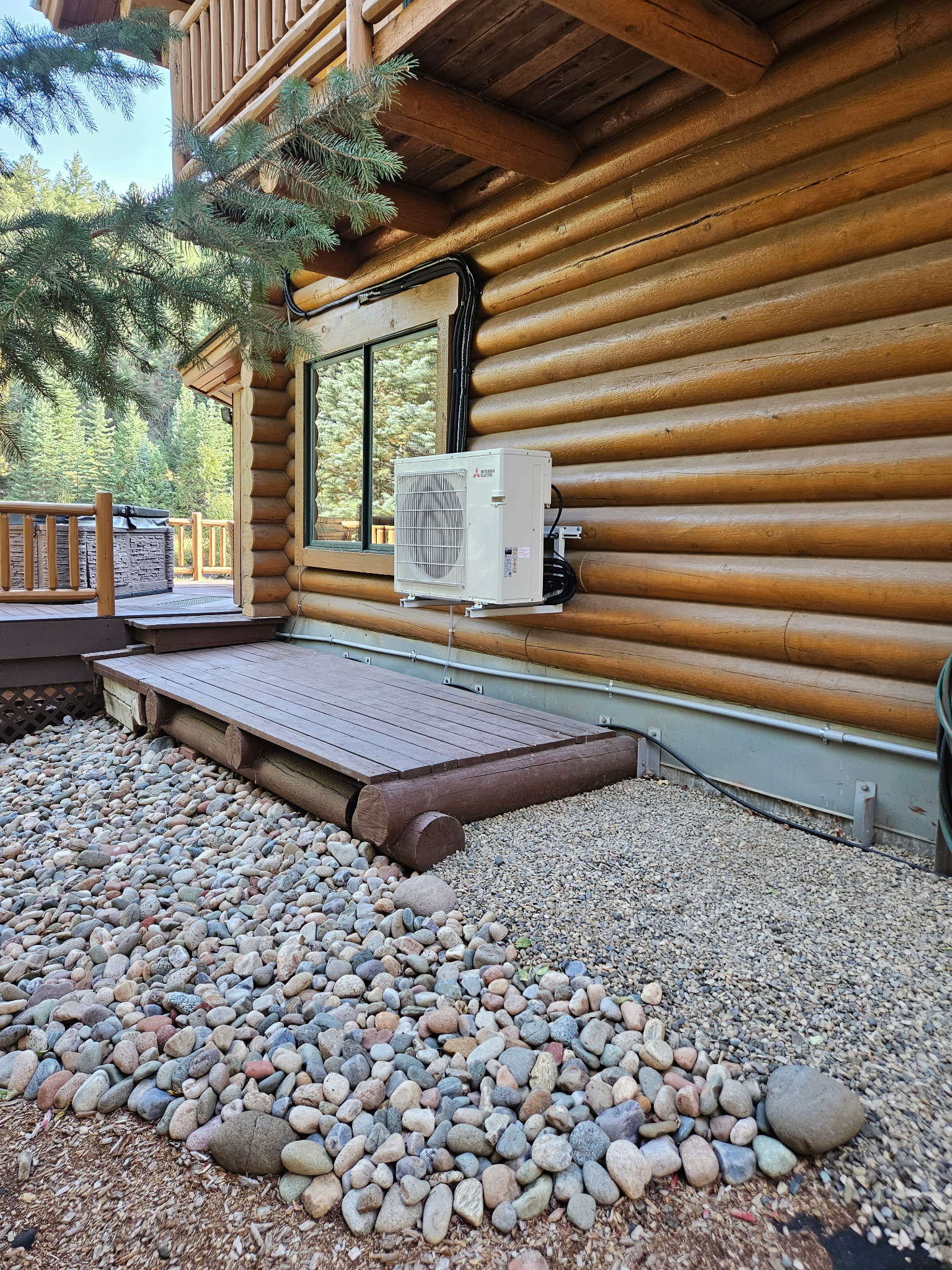 Mini-split outdoor unit mounted on the exterior of a log cabin above a river rock bed